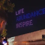 Personas observan un espectáculo nocturno de drones que forman palabras iluminadas en el cielo, leyendo ‘LIFE ABUNDANCE INSPIRE’, mientras algunos asistentes graban el evento con sus teléfonos.