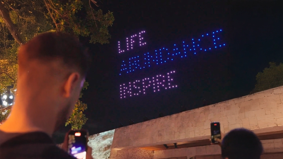 Personas observan un espectáculo nocturno de drones que forman palabras iluminadas en el cielo, leyendo ‘LIFE ABUNDANCE INSPIRE’, mientras algunos asistentes graban el evento con sus teléfonos.